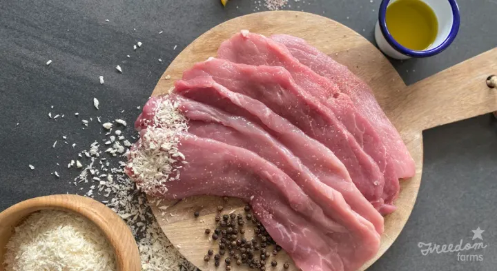 Image: Raw pork schnitzel on a chopping board, with peppercorns, panko crumbs and oil in the shot as well.